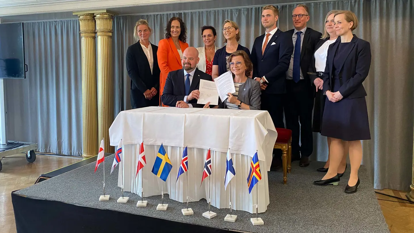 A group of people standing around a table with a white cloth where a man and a woman sits down holding a signed document.